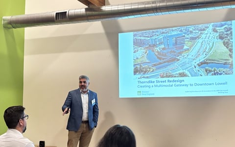 A person presenting and standing beside a screen displaying a slide titled "Thorndike Street Redesign: Creating a Multimodal Gateway to Downtown Lowell."