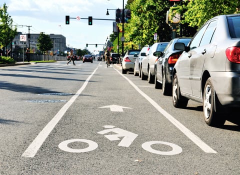 Cars parked beside a bike lane, with pedestrians crossing a crosswalk in the background.