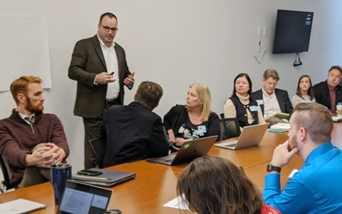 Dave Mulholland is holding a pen and standing before a large writing pad  leading a workshop with participants sitting around a conference table.