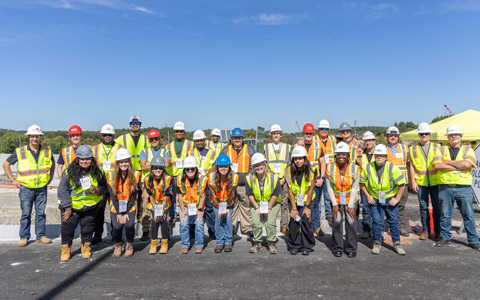 Group of construction workers and students in safety gear standing together in a line at a construction site.
