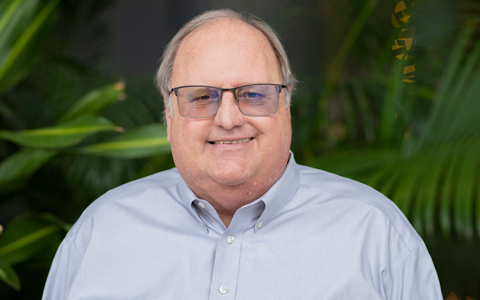 Neale Stralow wearing a gray shirt in front of palm leaves.