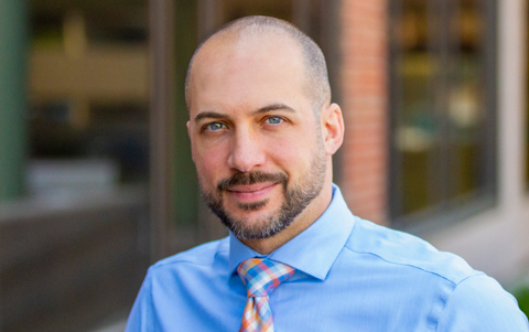 Headshot of Nick Scenna, a blue-eyed, bearded man in a blue dress shirt and tie.
