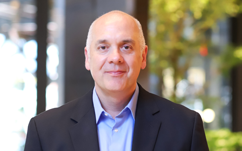 Joe wearing a navy blazer and blue shirt in front of a blurred background.