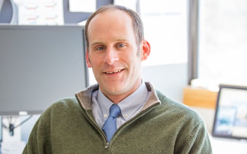Peter Smiar seated in an office, wearing a sweater over a collared shirt. The background includes a window and a computer monitor with various notes attached.