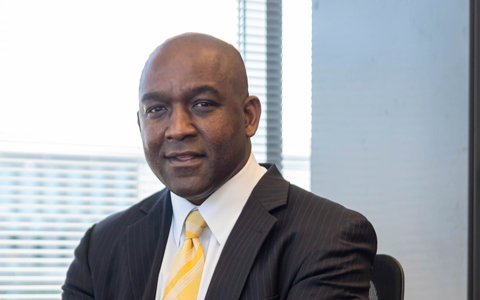 Reggie Scales sits at his desk in a black sports coat and yellow tie.