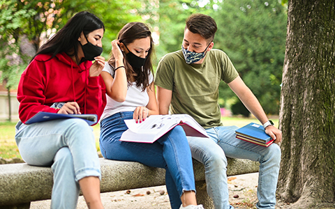 Three college students wearing masks on sitting on a bench while looking at a book together.