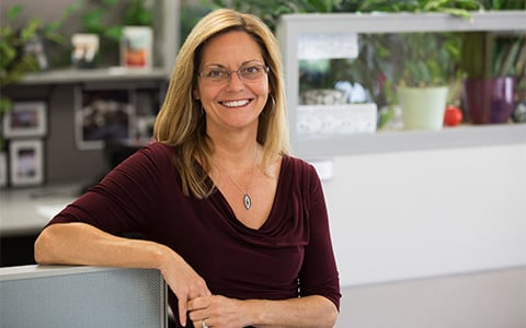 A woman with glasses standing in an office with plants behind her.