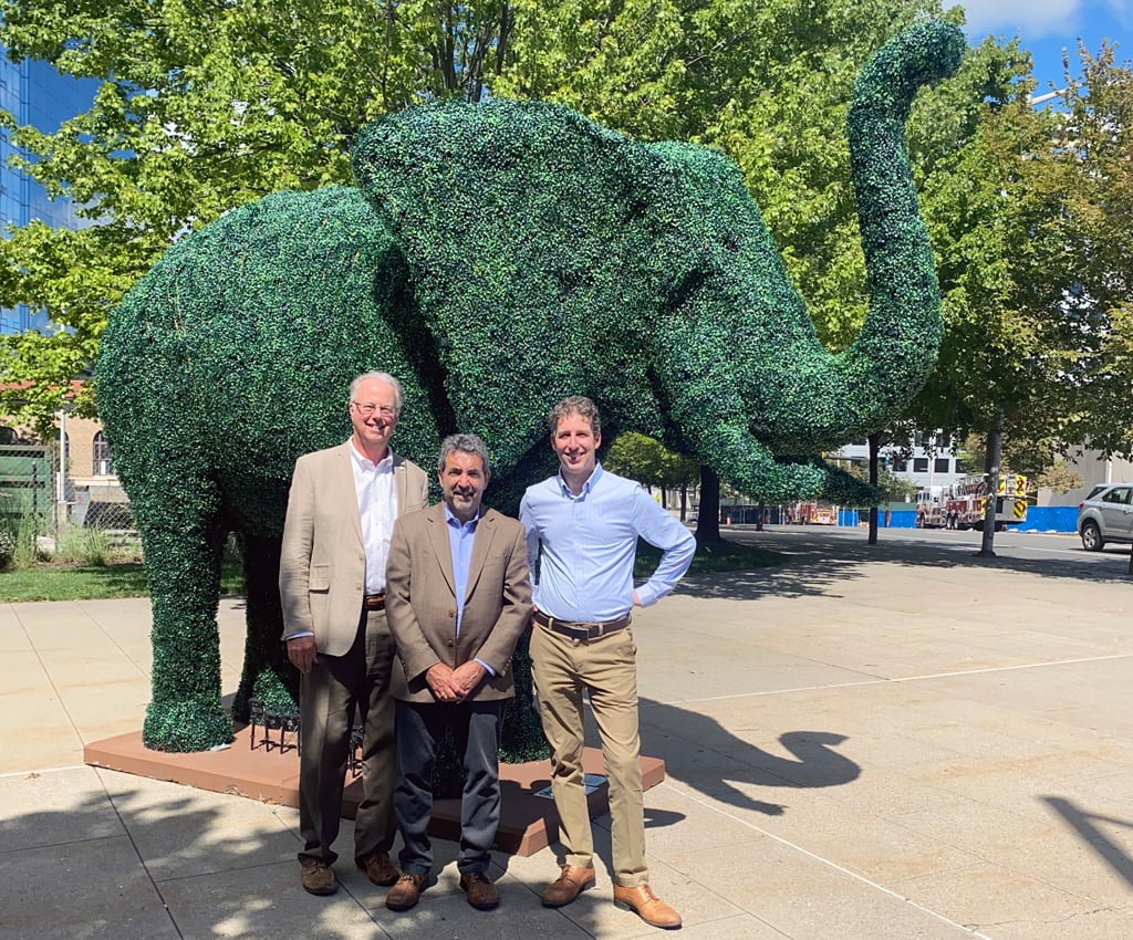 Three men stand in front of a green, elephant topiary on a sidewalk.