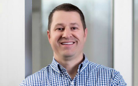 Stephen Quina leans against wall in blue checkered shirt.