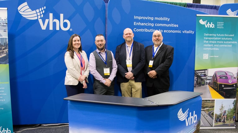 A group of four people stand in front of a VHB booth at a conference. The booth displays images of transportation and city planning projects.  