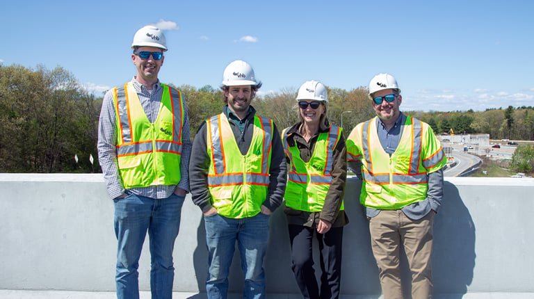 Four people are standing on a paved surface in front of a concrete barrier. They are wearing safety vests and hard hats with the VHB logo.  
