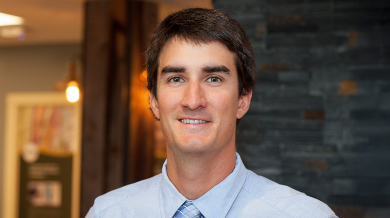 Person smiling in an office environment with a casual blue shirt and tie, standing in front of a rustic wooden beam and a dark stone column.