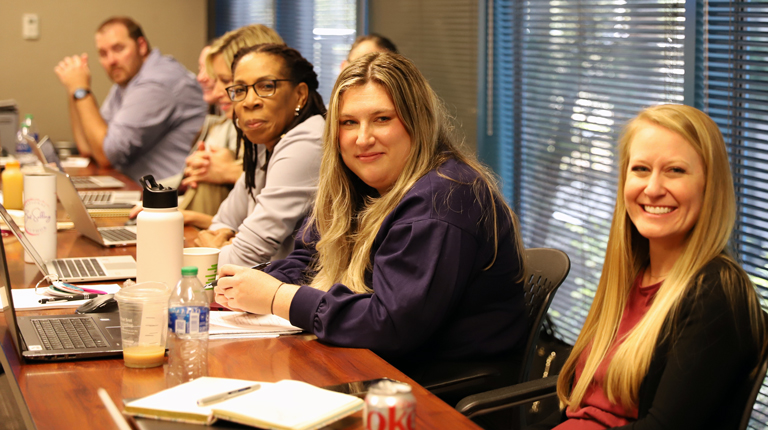 Alex DeYoung smiles in an office setting at a conference table with colleagues