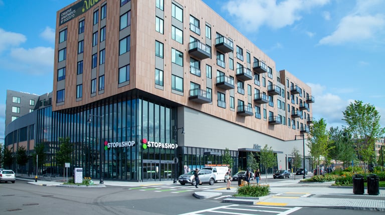 Modern urban building housing a Stop & Shop store on the ground floor, with a mix of glass and wooden facade, set against a backdrop of a clear blue sky with scattered clouds.