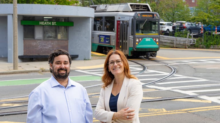 Two individuals smiling in front of a Heath Street station with a moving tram labeled 'E Line'.