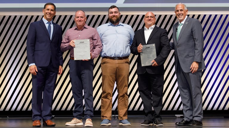 Five individuals are standing on a stage at a professional event, each holding a certificate, smiling, and dressed in business attire. 