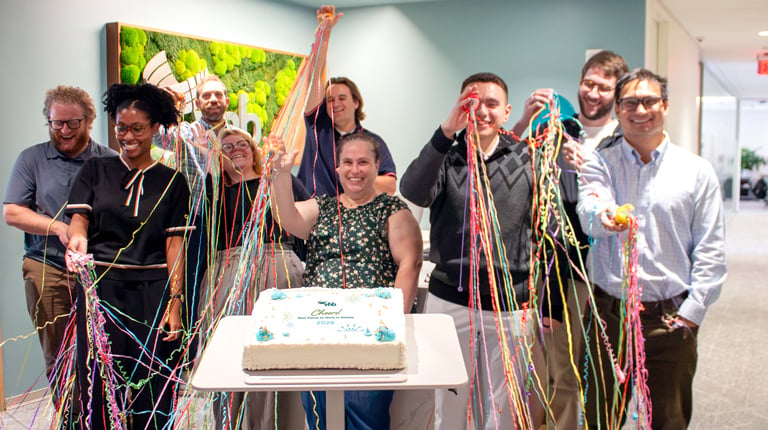 Employees stand around a celebration cake in a VHB office while launching confetti streamers.