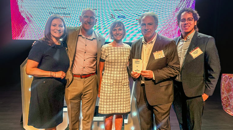 A group of five people smiling in front of a sign that reads "Corporate Citizenship Awards" from the Boston Business Journal. They are dressed in formal attire.