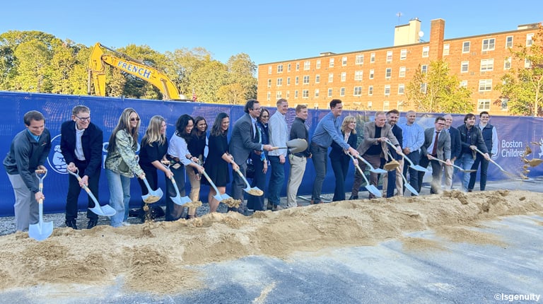 Group breaks ground with shovels at a construction site, with an excavator and Boston Children’s banner behind them.
