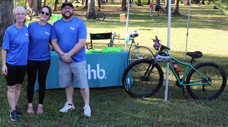 Three smiling VHBers standing in front of a teal VHB tent at the Bike 5 event.
