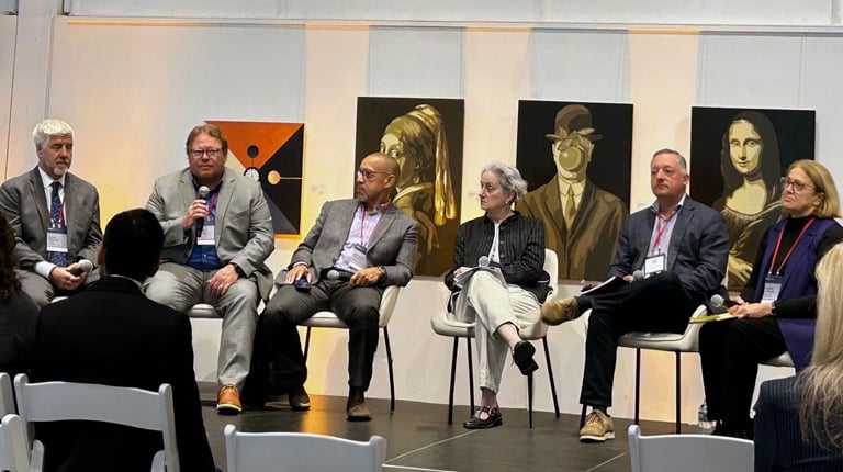 Panel of six speakers seated onstage at a conference. The audience is visible in the foreground, listening attentively.