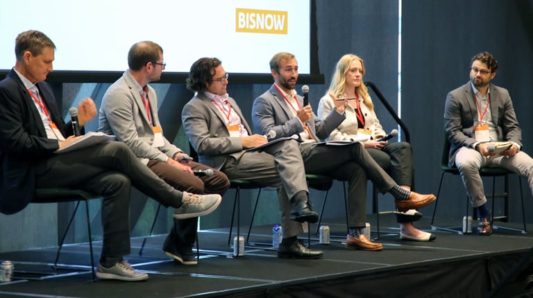 A panel of six individuals seated on stage at a Bisnow event. They are engaged in a discussion, each with a microphone.