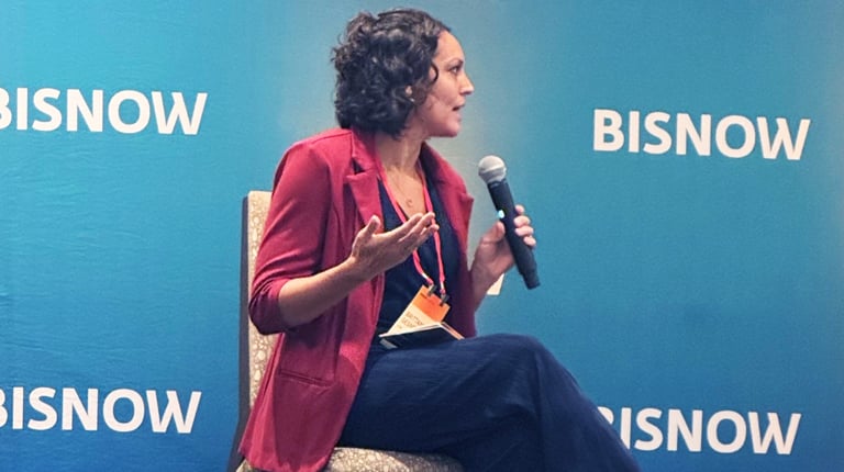 A person in a red blazer and dark pants sitting on a stage, speaking into a handheld microphone at a BISNOW event.
