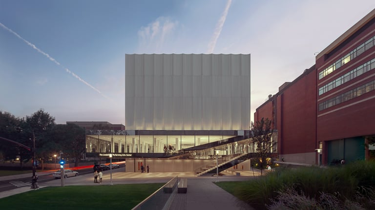 A dimly lit large building with a blue and purple sunset behind it and students walking through the site.