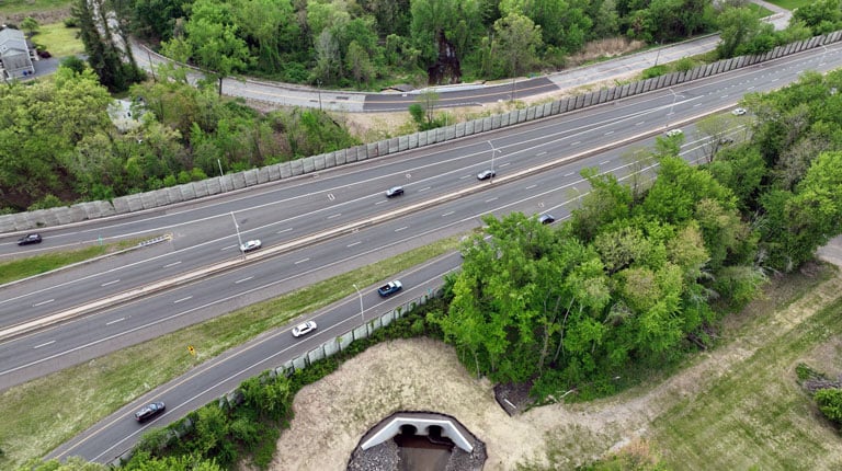 Aerial view of a highway with four lanes showing moderate traffic, surrounded by lush greenery and a small tunnel passing under one side of the road.