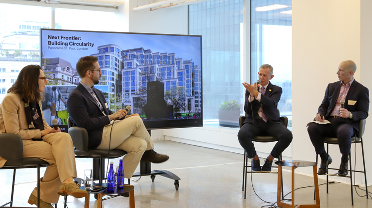 A panel discussion featuring 4 speakers seated in front of a screen displaying building images.