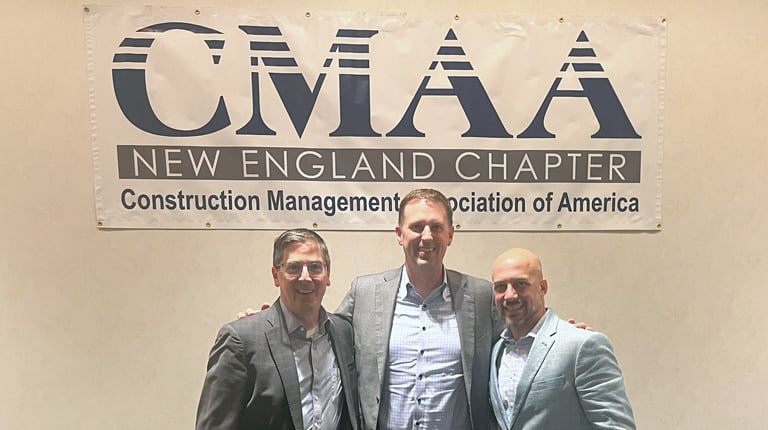 Three individuals stand smiling in front of a banner that reads "CMAA New England Chapter, Construction Management Association of America."