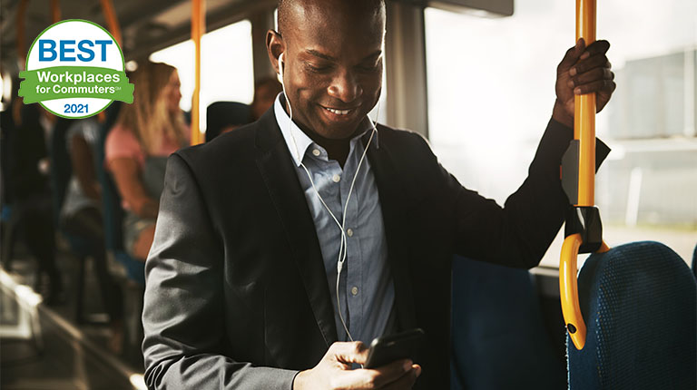 A young worker wearing a button down shirt and jacket with earphones listens to music on his phone while holding standing and holding a rail in a bus