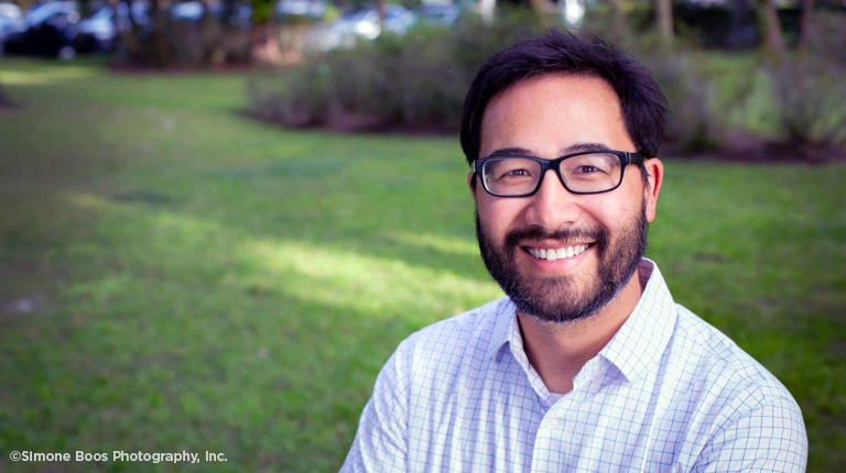 A business casual headshot of a man with dark hair, glasses and a beard featured outside in a garden setting.