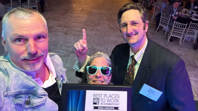 Three individuals on a stage taking a selfie holding a Best Places to Work plaque with a crowd in the background. 