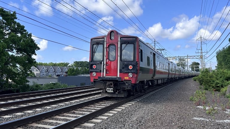 A red and silver Metro-North Railroad train travels along a track on a clear day with scattered clouds. Trees and houses are visible in the background.
