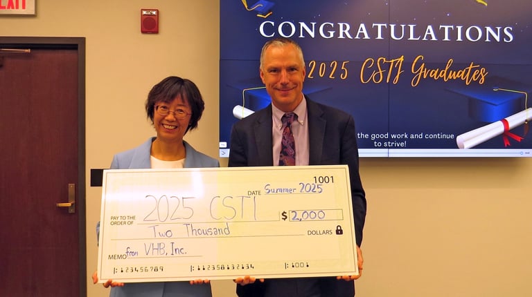 Two individuals holding a large ceremonial check from VHB, Inc., celebrating CSTI Graduates under a "CONGRATULATIONS" banner