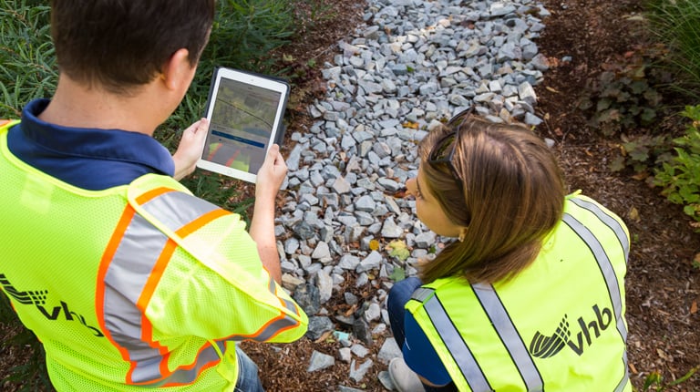 Two individuals in high-visibility vests are crouched over a tablet by a gravel path, examining a digital map.