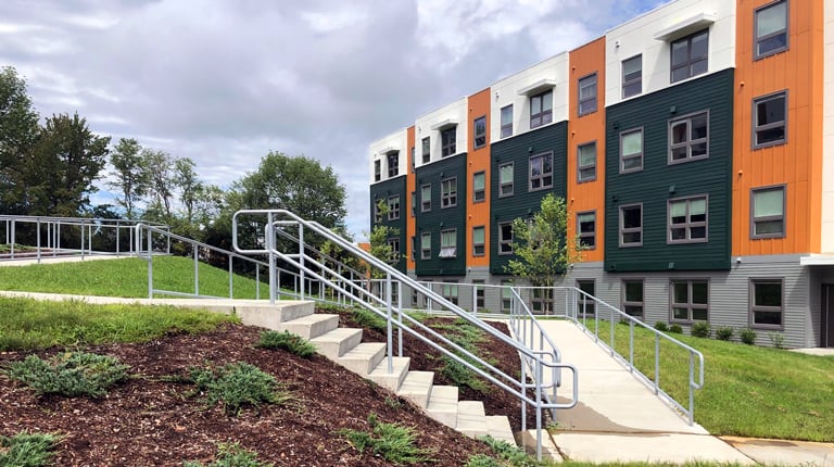 A white, green, and orange student housing building is bordered by accessible paths and green space. 