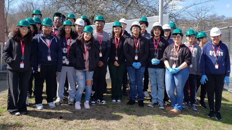 Group of student volunteers wearing green hard hats and blue gloves standing outdoors for a community clean-up event.