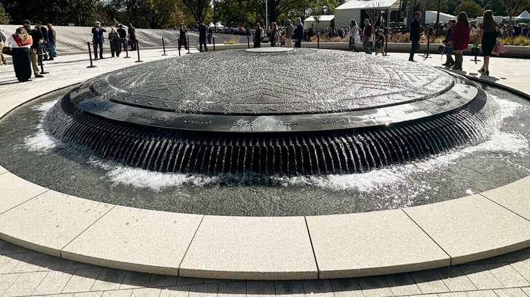 A large bronze shield fountain is at the center of the memorial, surrounded by visitors and blue skies.