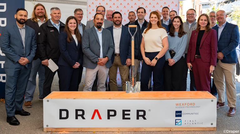 A group of individuals stands smiling in front of a backdrop displaying logos, including Draper, Wexford Science & Technology, GMH Communities, and SmartLabs, at a groundbreaking event with a silver shovel placed prominently.