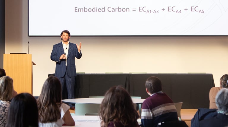 A person is giving a presentation to an audience in a conference room. The slide on the screen contains text on topics such as embodied carbon and related formulas.  