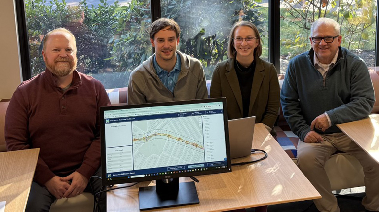 Four people smiling in an office setting beside a laptop computer.