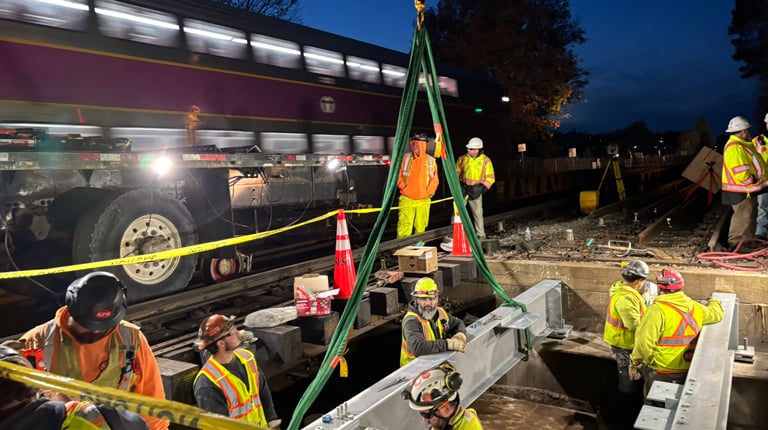 Workers in high-visibility clothing install large equipment near a commuter train during nighttime hours.