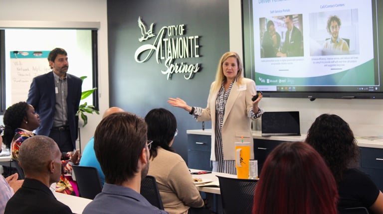 Two presenters at the front of the room in front of smart screen discussing a PowerPoint. 