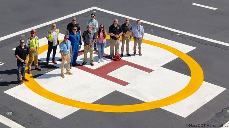 Aerial image of the project team smiling on a helipad with the hospital behind them.