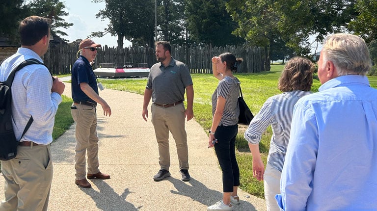 A group of people stand on a paved pathway at Jamestown Island, discussing site conditions during an outdoor project visit.