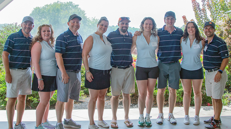 1.	A group of eight individuals standing together inside a patio area, all dressed in navy blue and white golf attire, with a background of colorful plants and trees bathed in soft daylight.