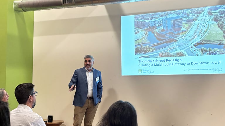 A person presenting and standing beside a screen displaying a slide titled "Thorndike Street Redesign: Creating a Multimodal Gateway to Downtown Lowell."