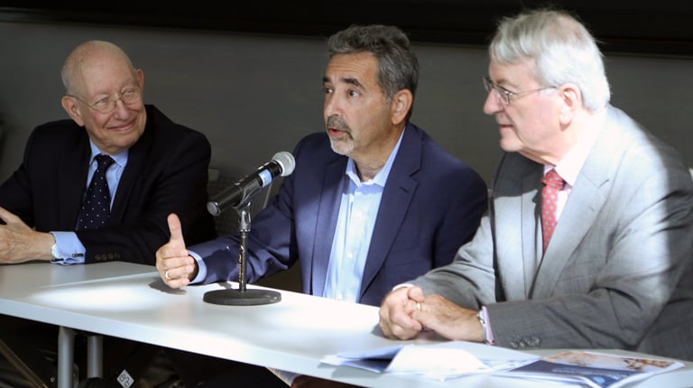 Three individuals participating in a panel discussion. The person in the center is speaking into a microphone, with another panelist on each side.